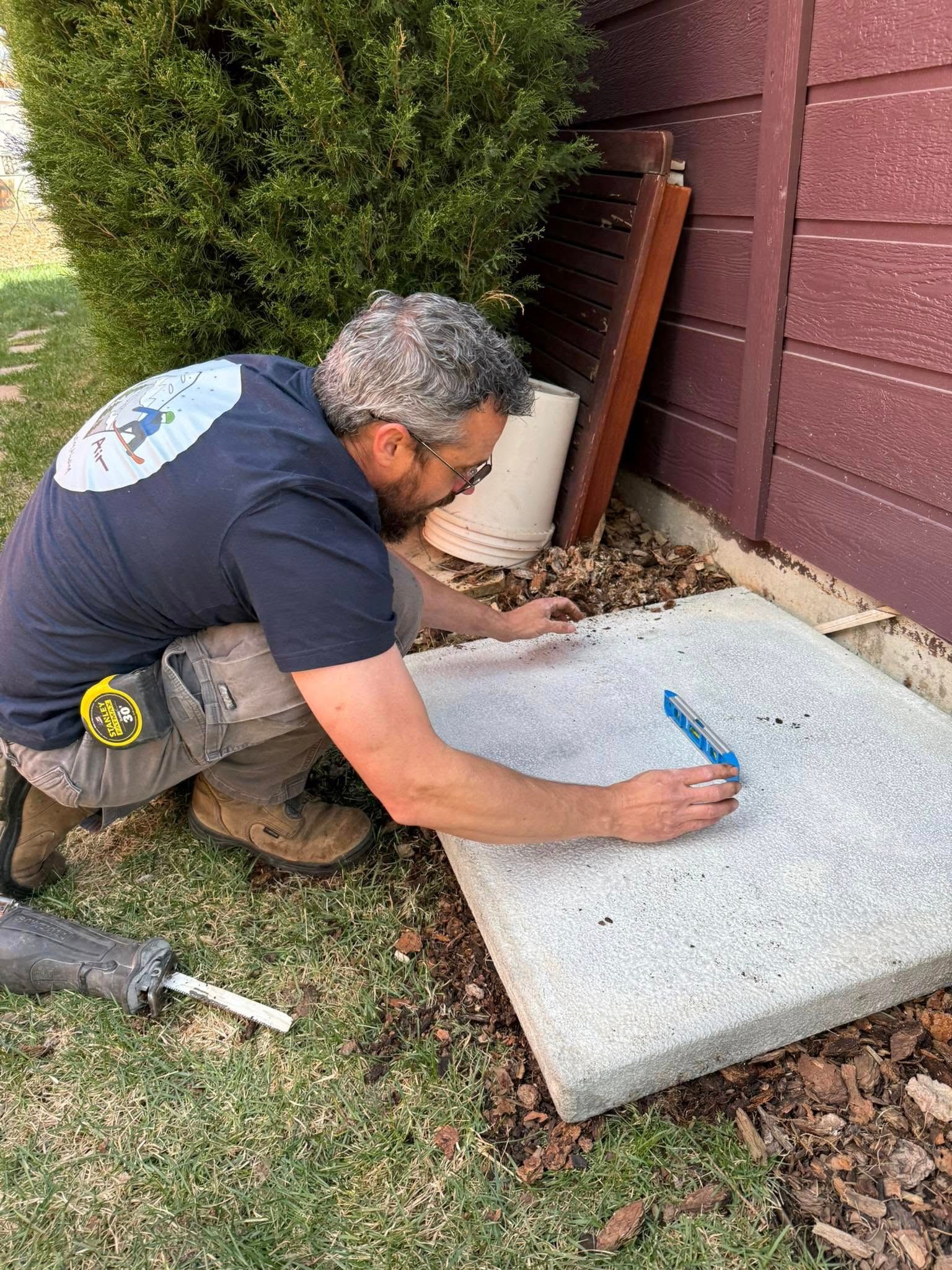 Man kneeling outdoors, marking a white concrete slab with a blue pencil beside a house wall