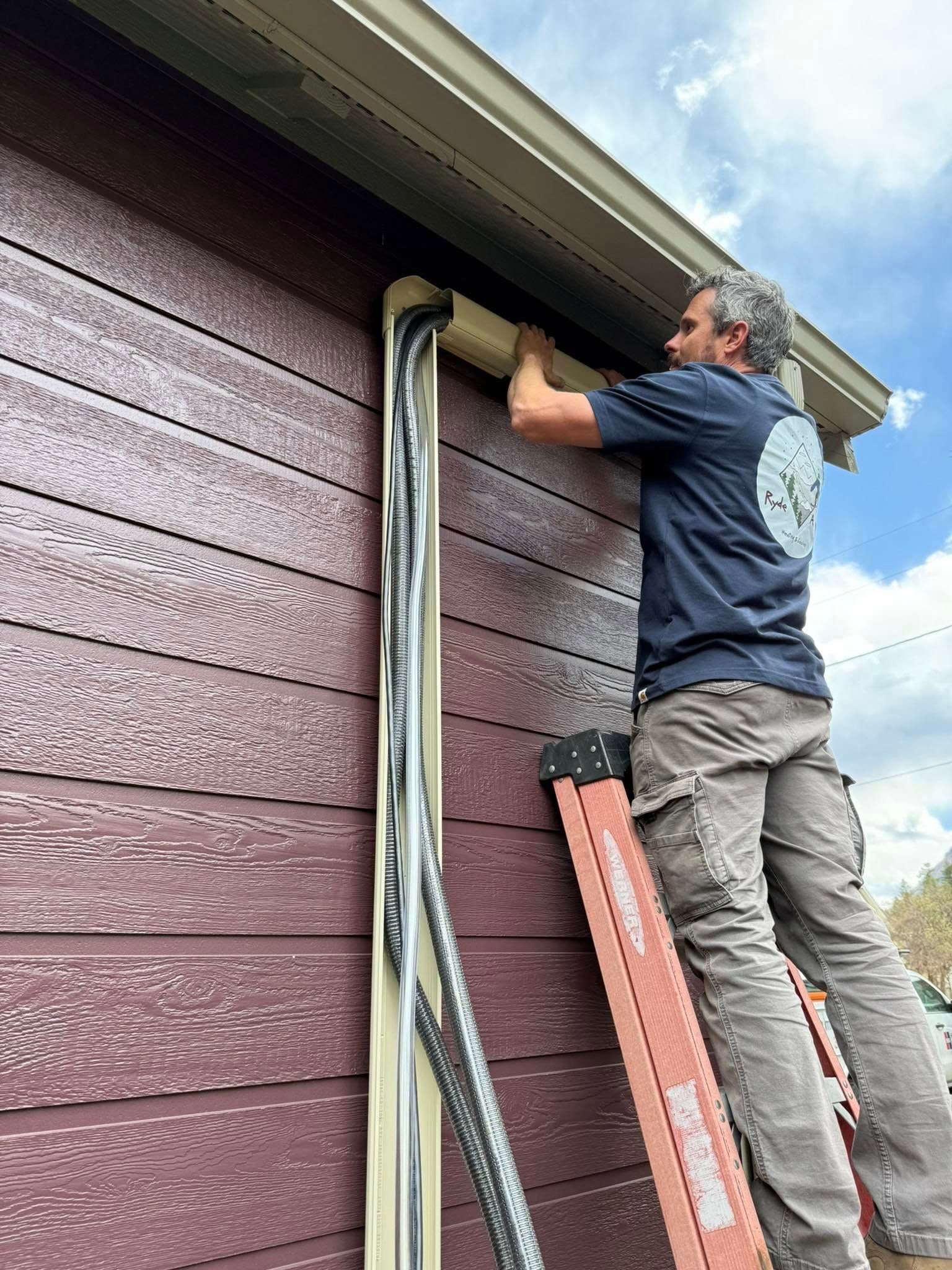 Worker on a ladder repairing roof trim on a maroon house.