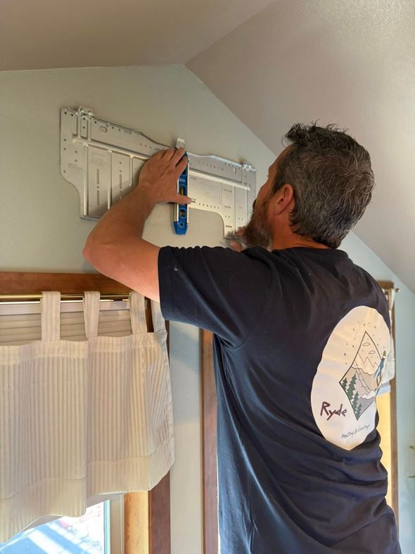 Person mounting a white wall shelf above a window with a blue drill in a bright room