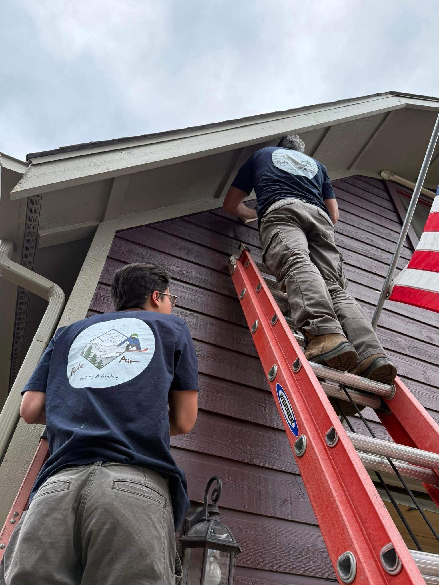 Two workers on a red ladder repairing a house roof and siding.
