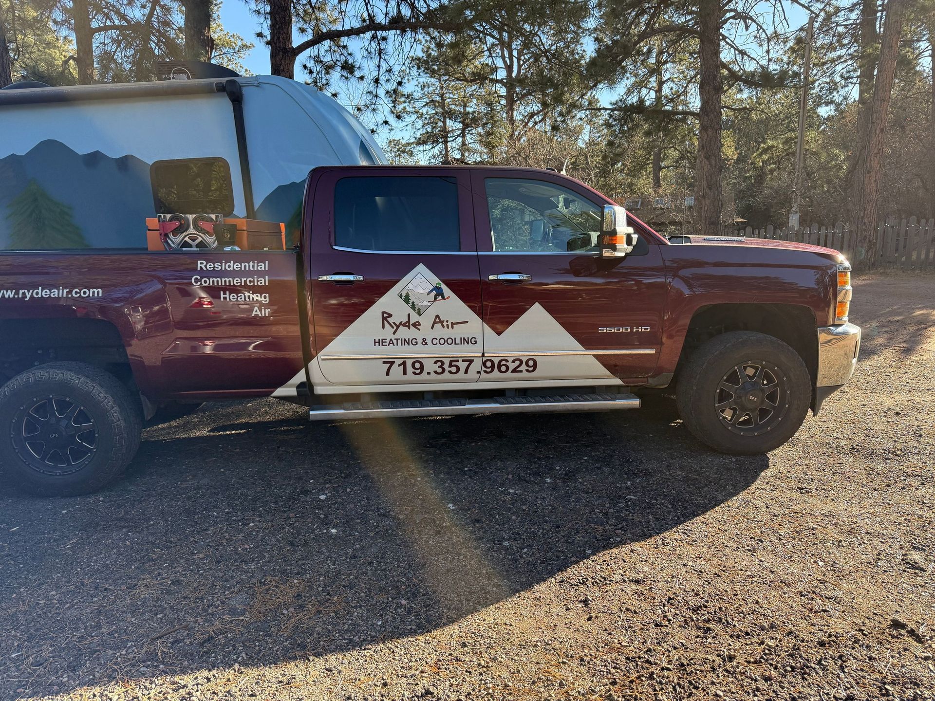 Maroon service truck with mountain-logo sign parked on gravel in a wooded area