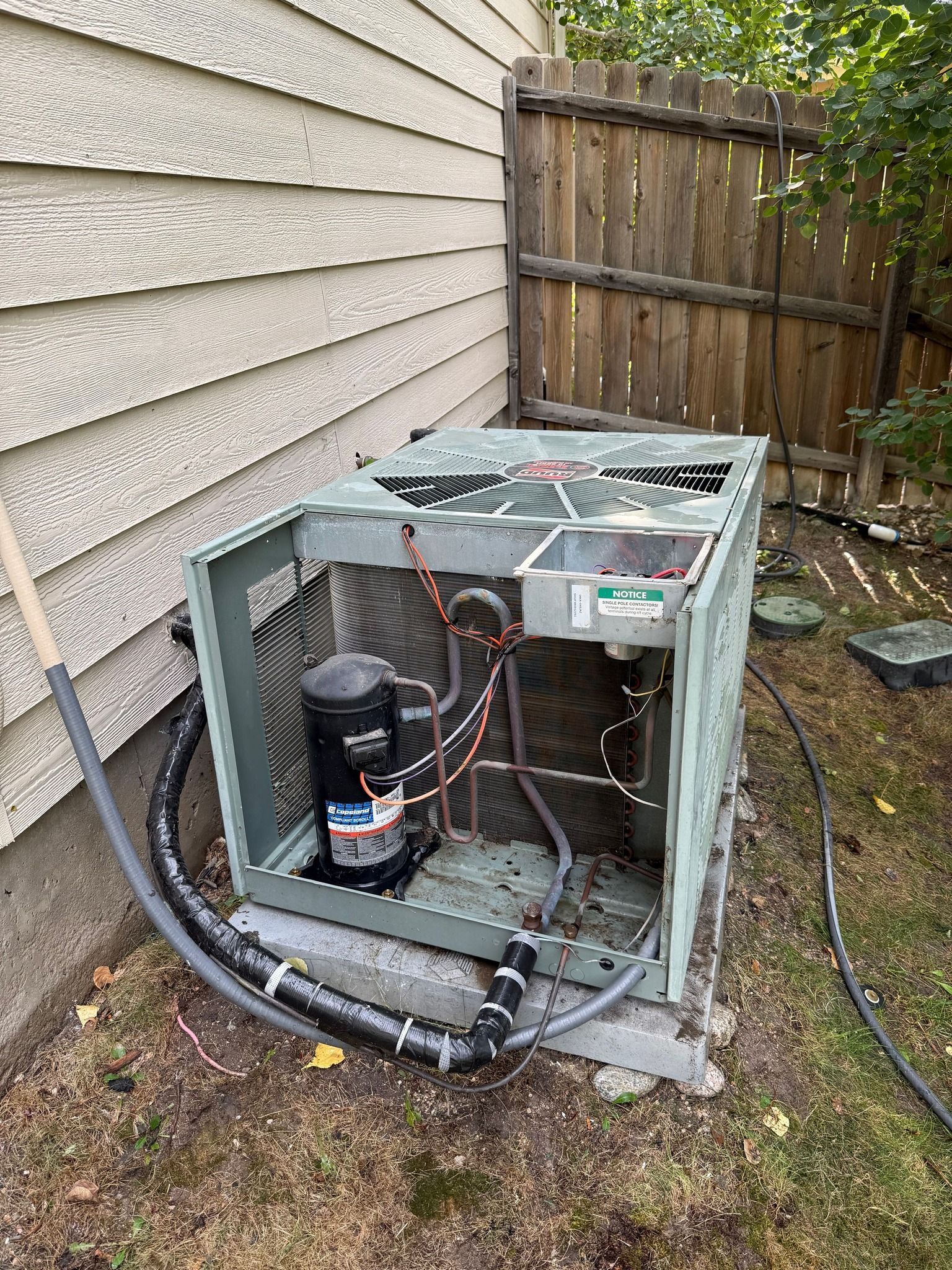 Outdoor HVAC unit with open side panel beside a house and wooden fence, showing exposed wiring and components.