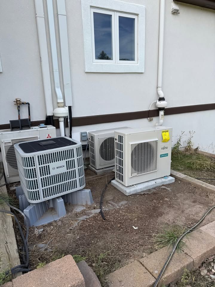 Two outdoor air conditioning units beside a white house wall and window, with exposed pipes and bare ground.