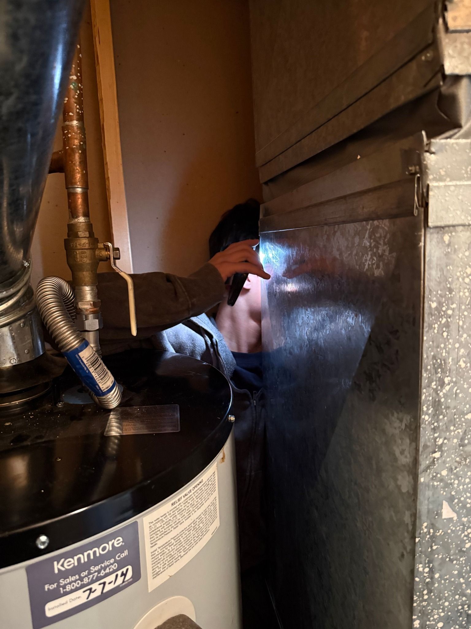 Technician inspecting HVAC unit with flashlight inside a metal duct in a cramped utility space