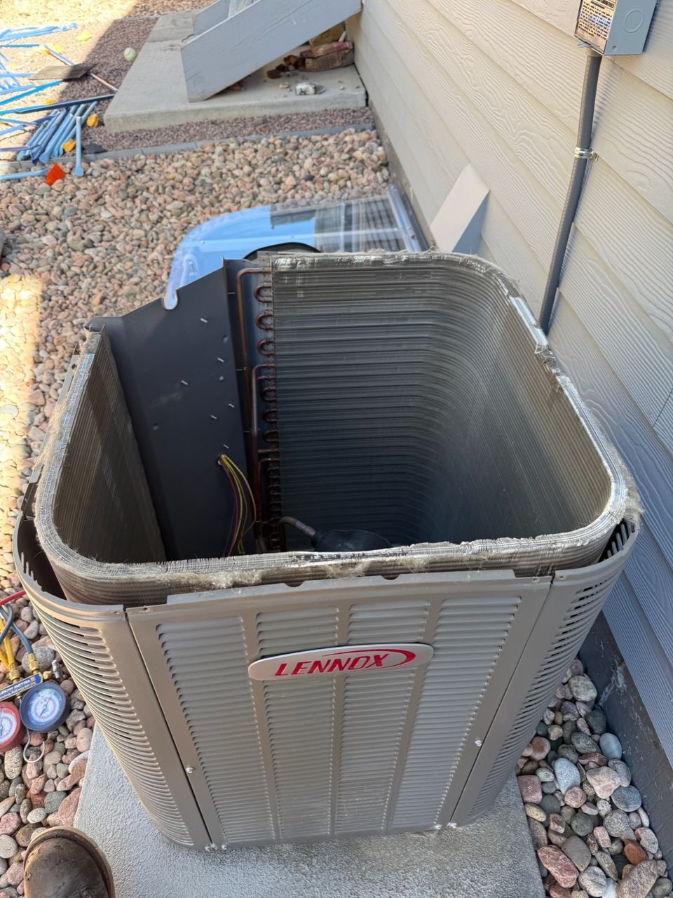 Open HVAC unit with damaged top panel beside a house wall and gravel ground