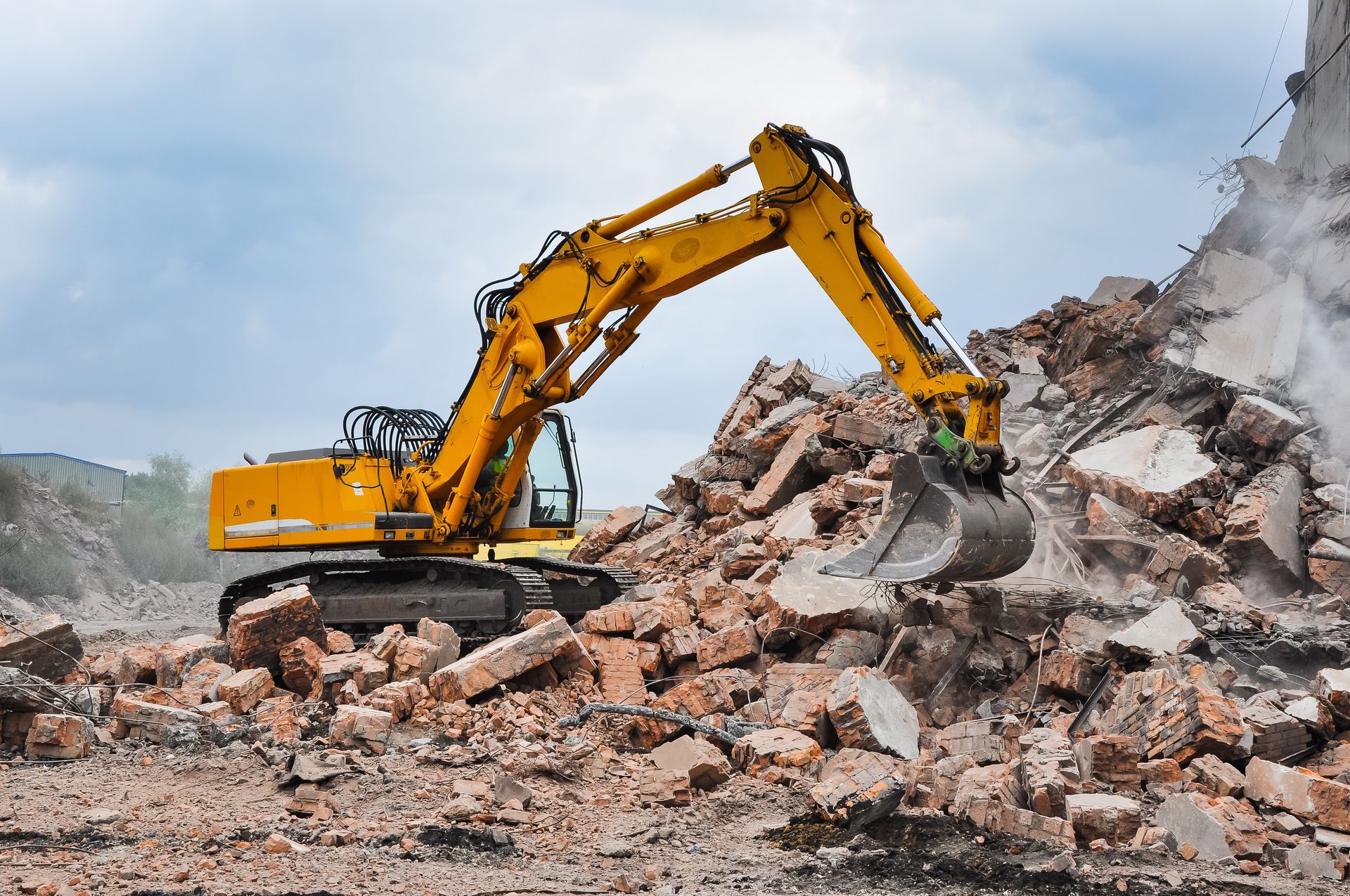Yellow excavator clearing rubble and debris at a demolition site under a cloudy sky.