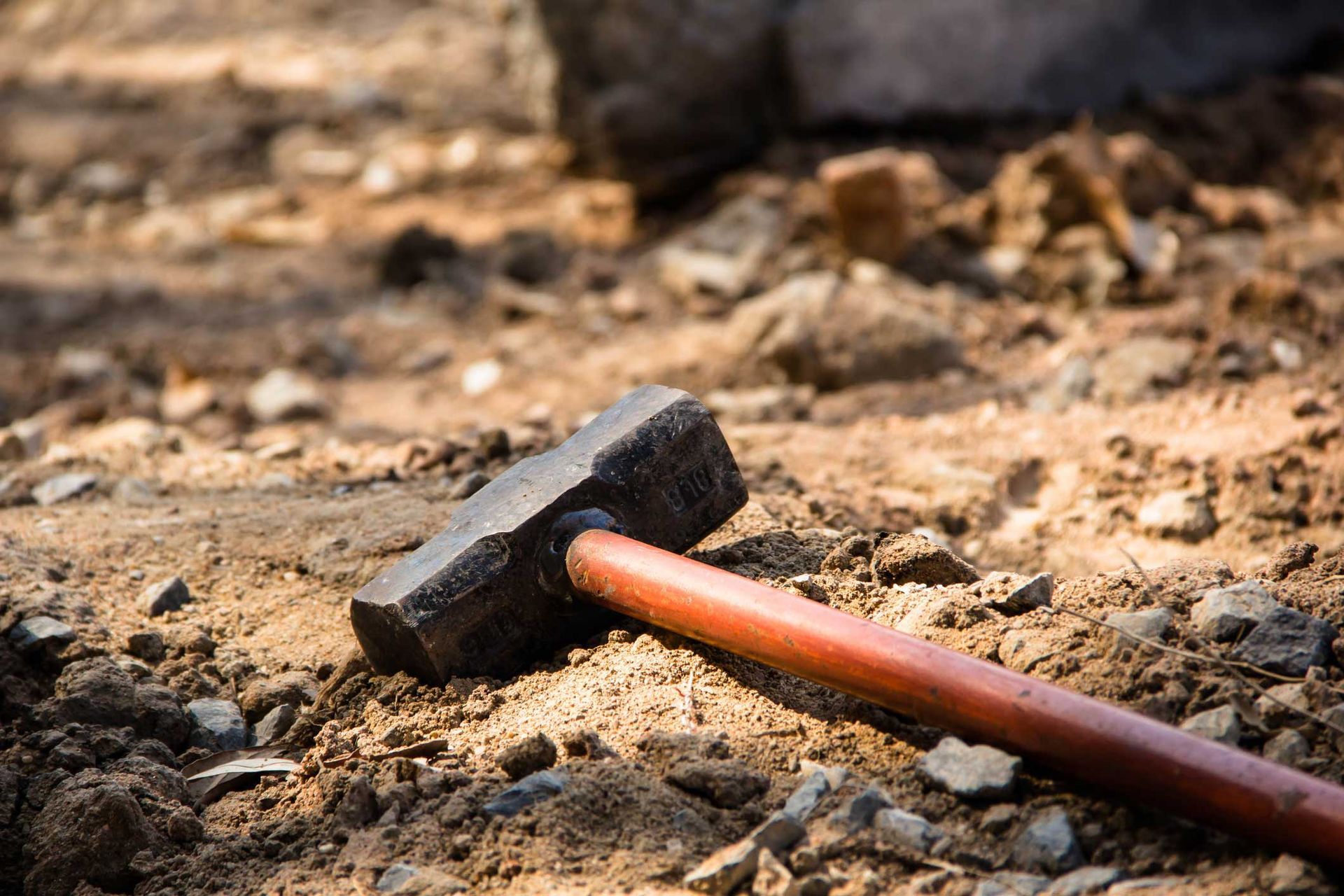 Red-handled sledgehammer on rocky dirt ground at construction site for demolition or building work.