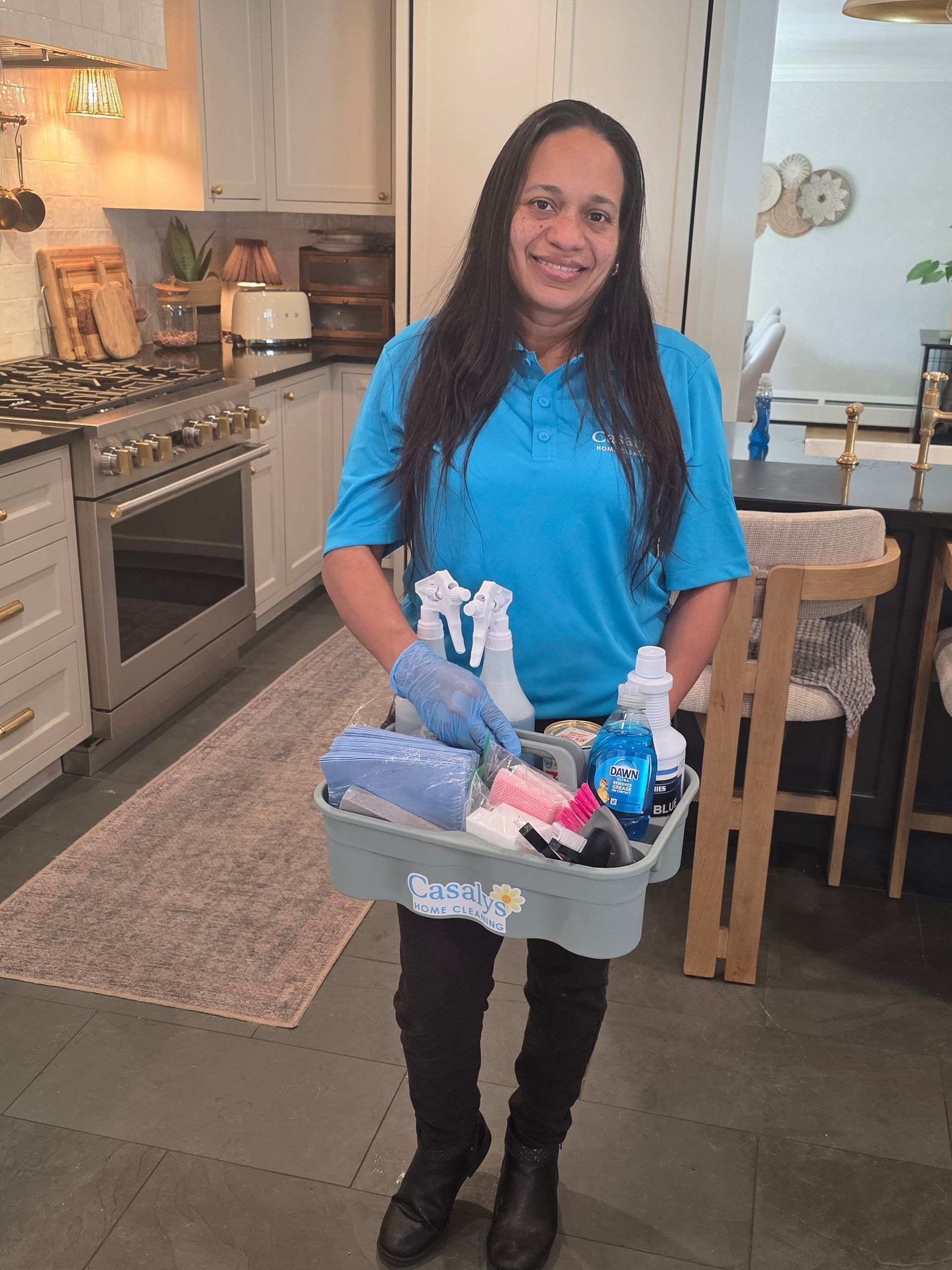 Woman standing in a kitchen holding a basket of cleaning supplies, wearing a blue shirt and black pants.
