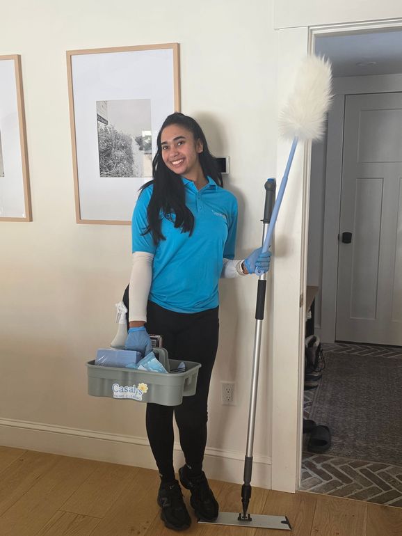 Woman in blue shirt holding cleaning supplies and a mop, standing indoors and smiling.