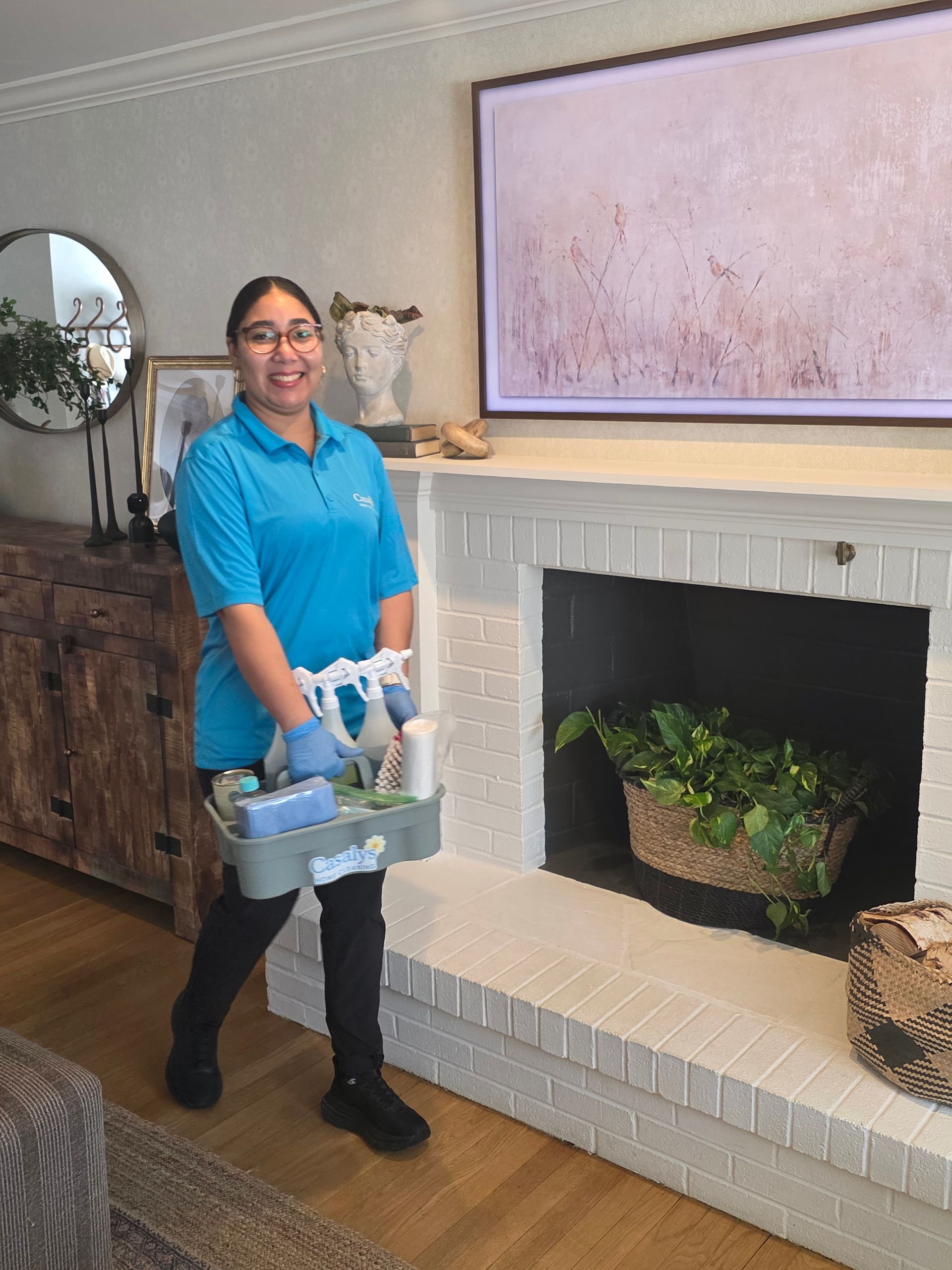 Person in a blue shirt holding cleaning supplies in a living room beside a white fireplace and framed art