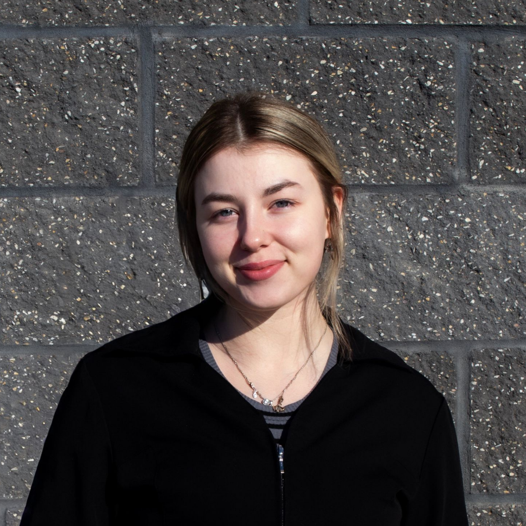 A woman in a black jacket is smiling in front of a brick wall.
