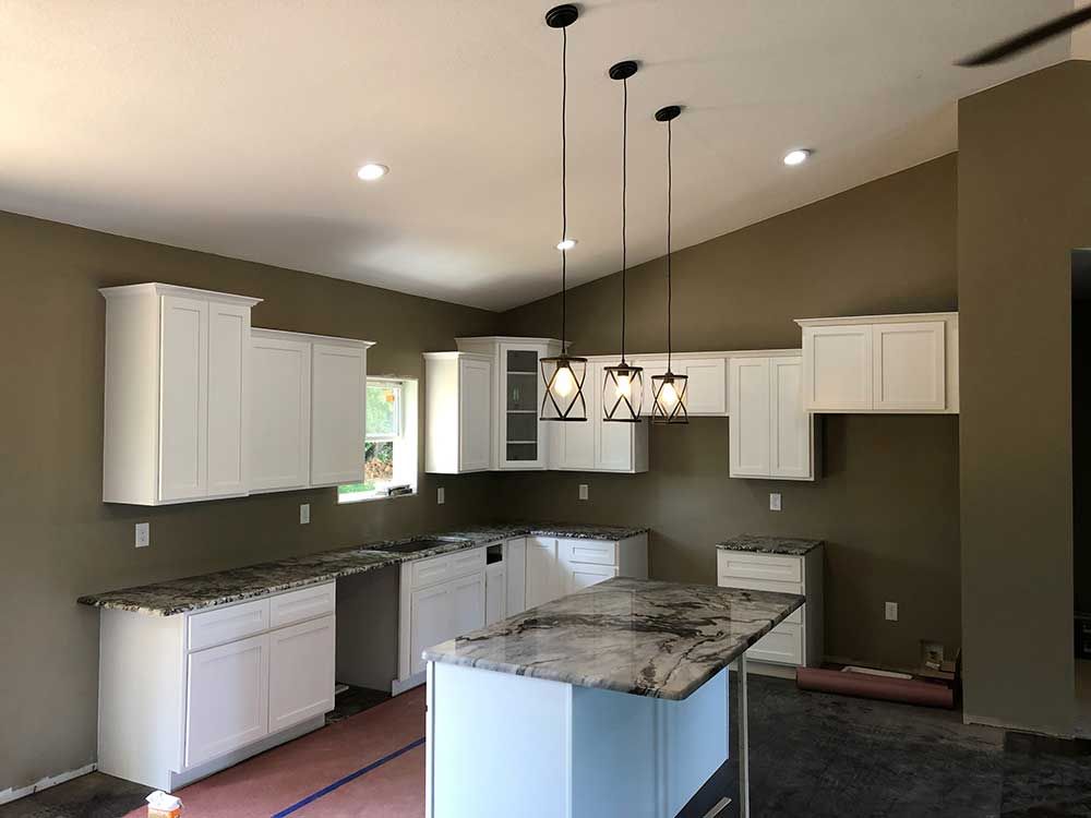 Empty kitchen with white cabinets, granite island, olive walls, and three pendant lights.