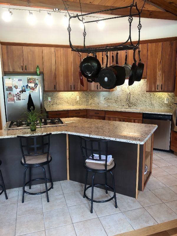 Kitchen with island, granite countertops, hanging pots, wood cabinets, and tiled floor.