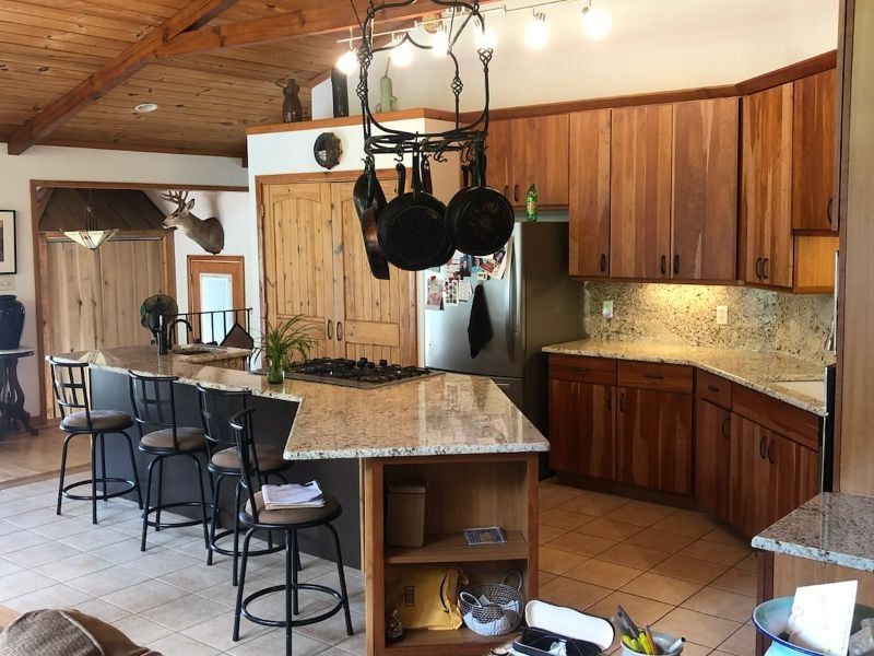 Warm kitchen with wood cabinets, granite island, hanging pots, bar stools, and tiled floor
