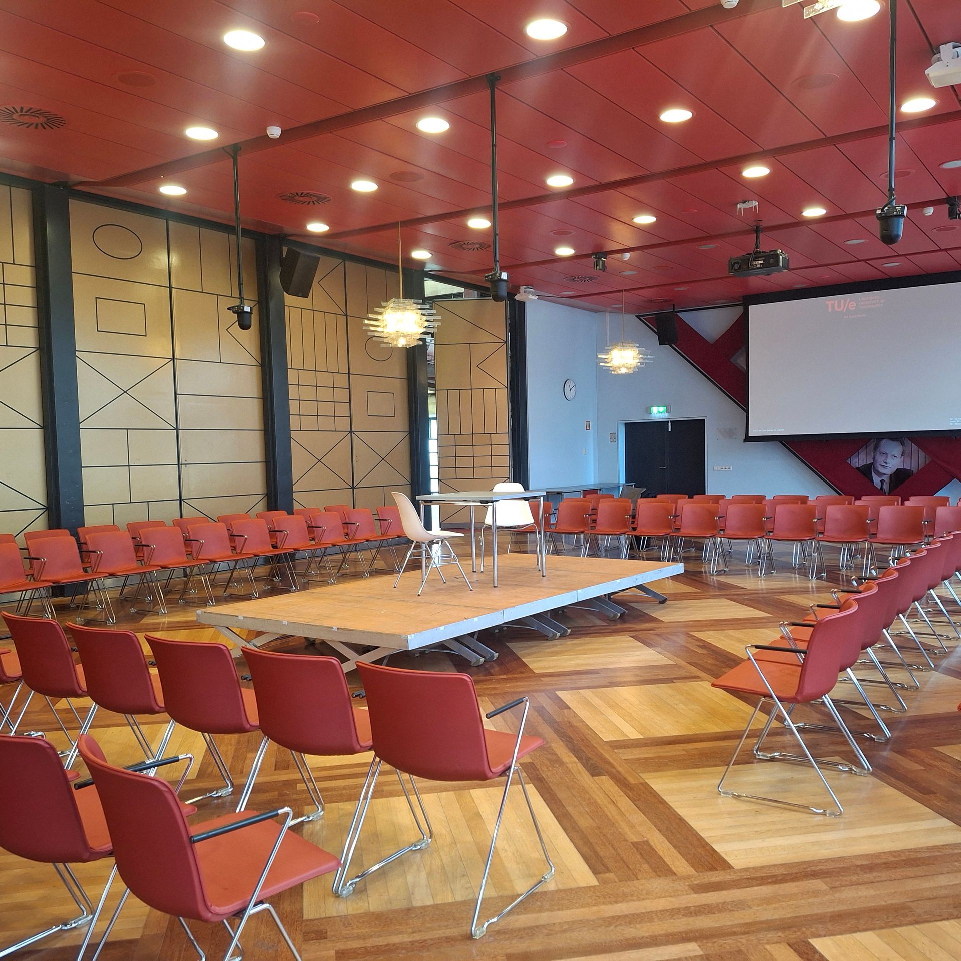 Vergaderruimte in rood thema met stoelen in een cirkel rond een klein podium. Projectiescherm zichtbaar.
