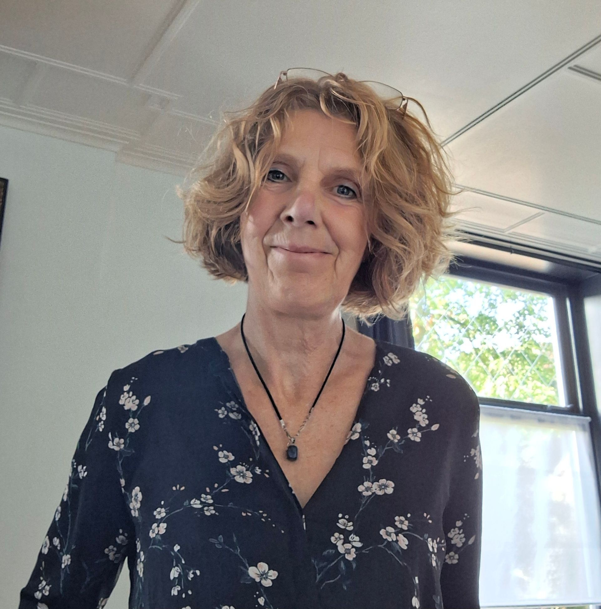 Lachende vrouw in een donkerblauwe blouse met bloemenprint staat binnen bij een raam en plafondpanelen.