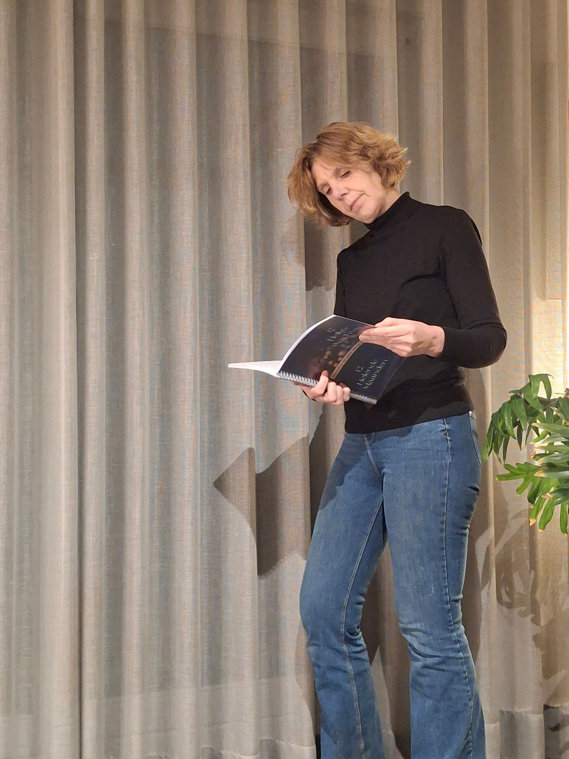 Een persoon in een zwarte top en spijkerbroek leest uit een boekje op een podium met grijze gordijnen en een potplant.