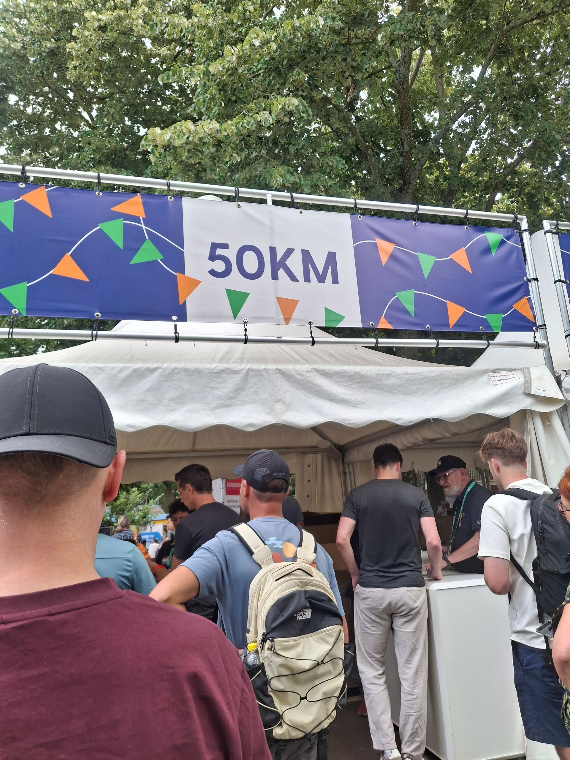 Een menigte verzamelde zich onder een tent bij een controlepunt van de 50 km-race, met een spandoek en bomen over de vloer.