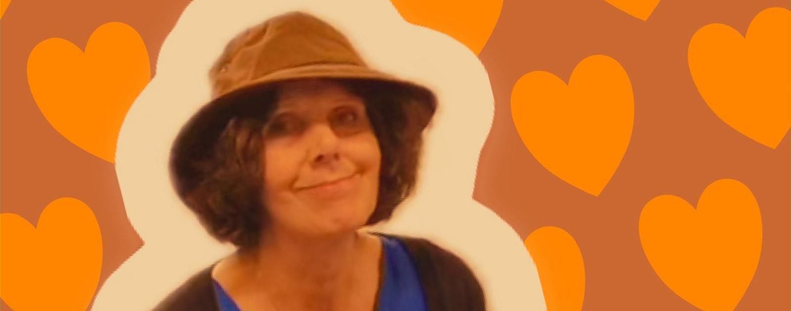 Een vrouw met een hoed lacht tegen een achtergrond van oranje hartjes.