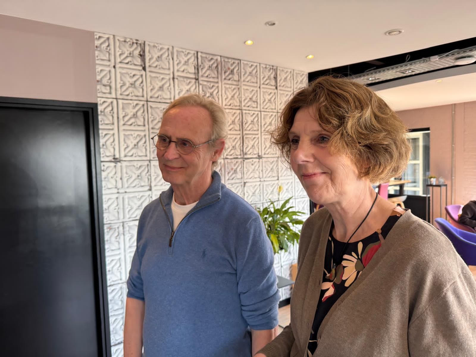 Twee personen staan binnen, vlakbij een zwarte deur en een muur met een patroon, en kijken niet in beeld.