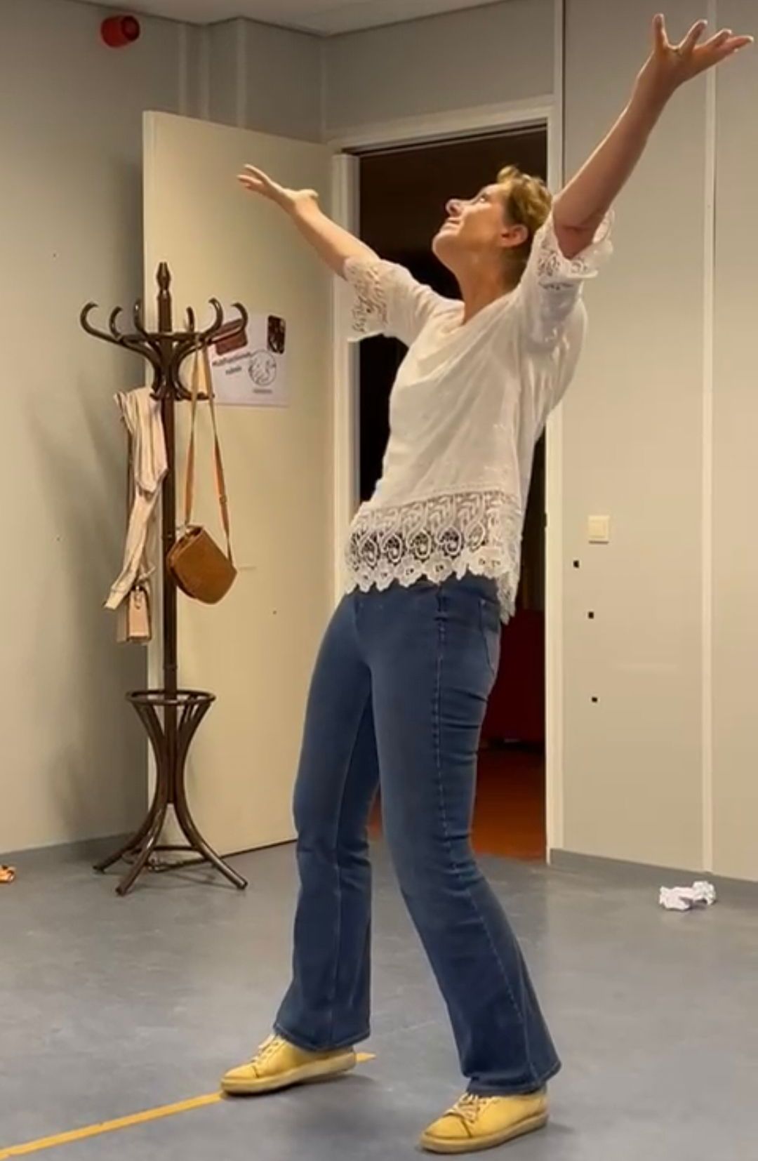 Vrouw in witte top en jeans met opgeheven armen in een kamer.