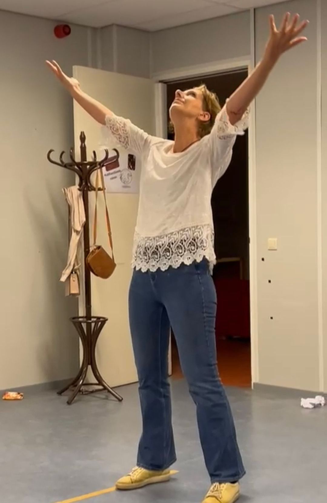 Vrouw met opgeheven armen in een kamer met een kapstok en een open deur.