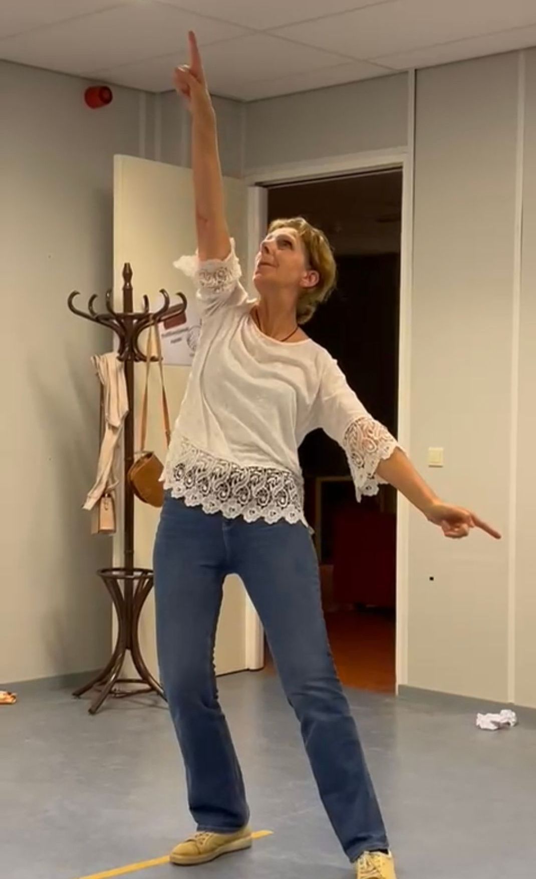 Vrouw wijst omhoog en naar rechts, in een danspose. Ze draagt ​​een spijkerbroek en een witte top. Binnen, bij een kapstok.