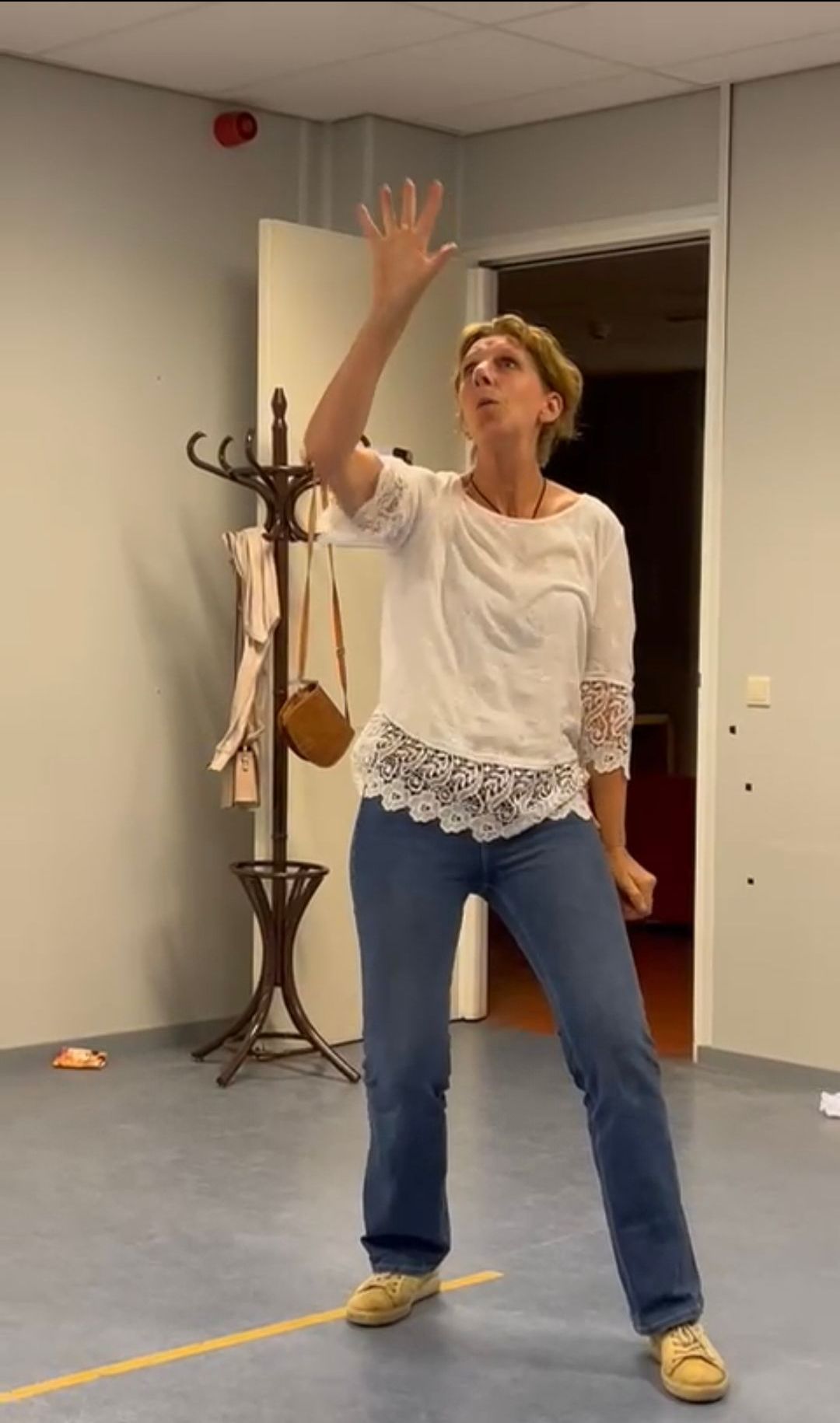 Vrouw in witte blouse en jeans met opgeheven arm en omhoogkijkende ogen in een kamer met een kapstok.