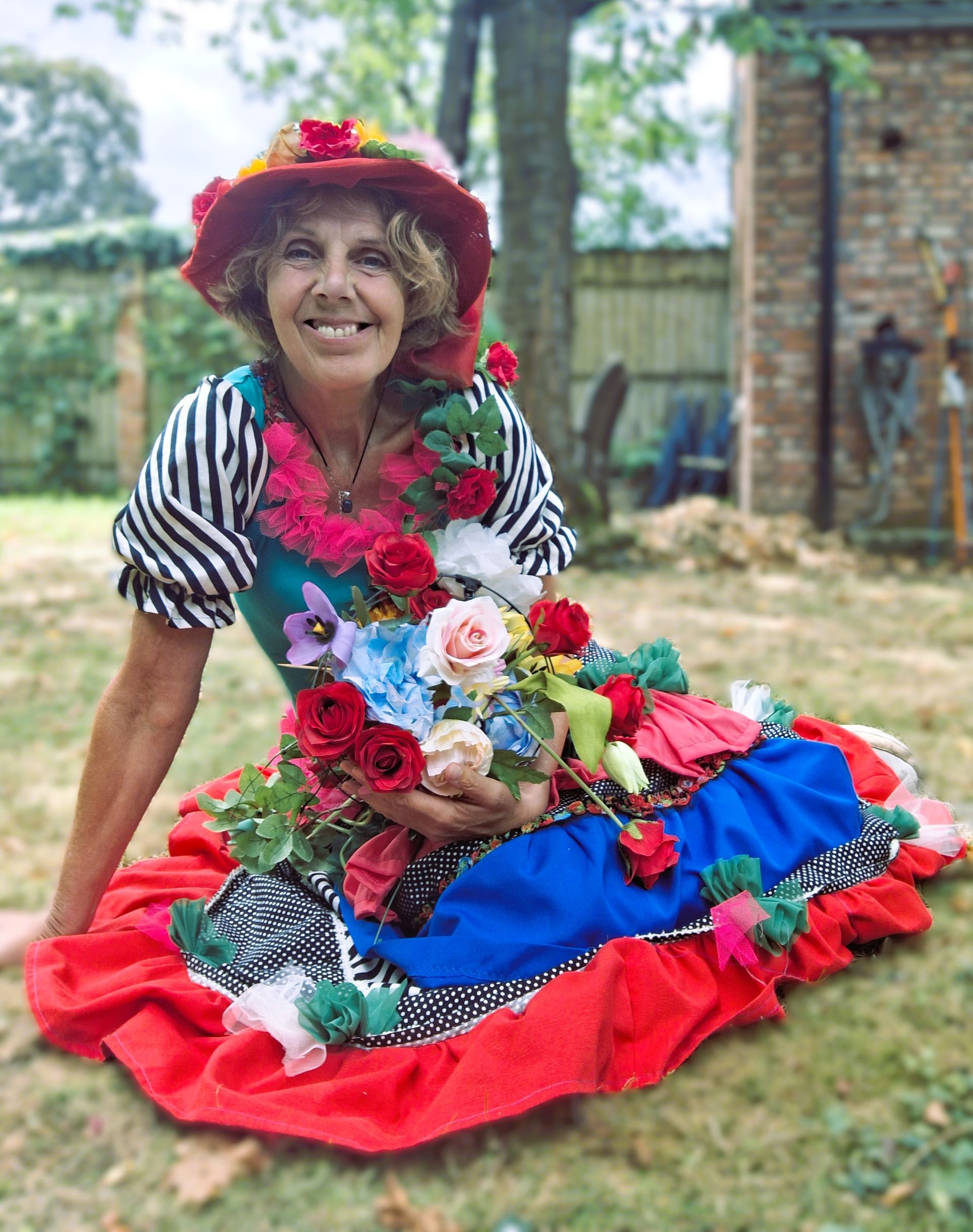 Een lachende vrouw met een rode hoed en een kleurrijke jurk zit buiten en houdt een boeket bloemen vast. 