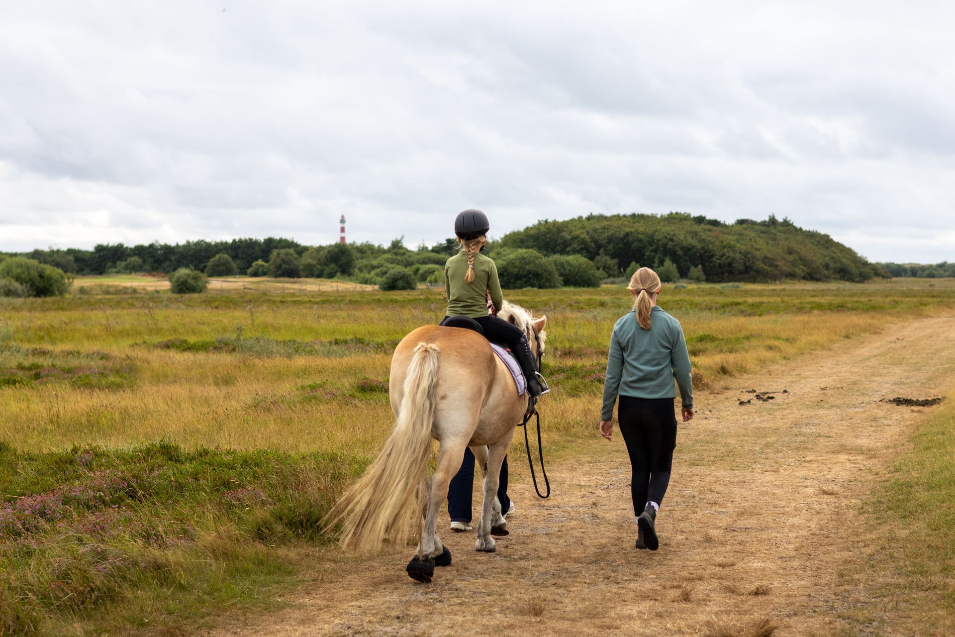 Een persoon die met een ruiter op een paard over een zandpad door open grasland loopt.