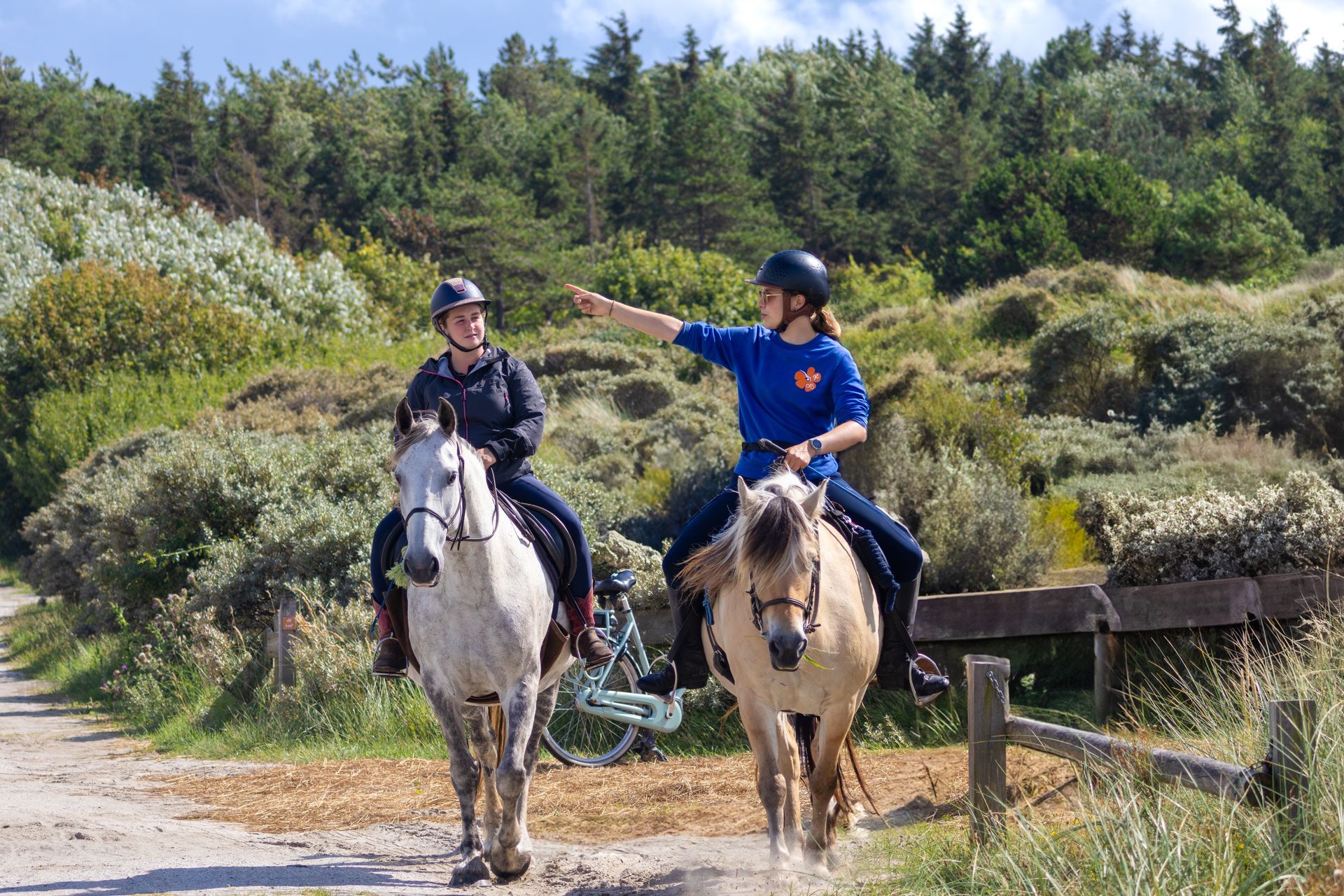 Twee mensen te paard rijden over een zandpad, één wijst naar rechts. Groen gebladerte en bomen op de achtergrond.