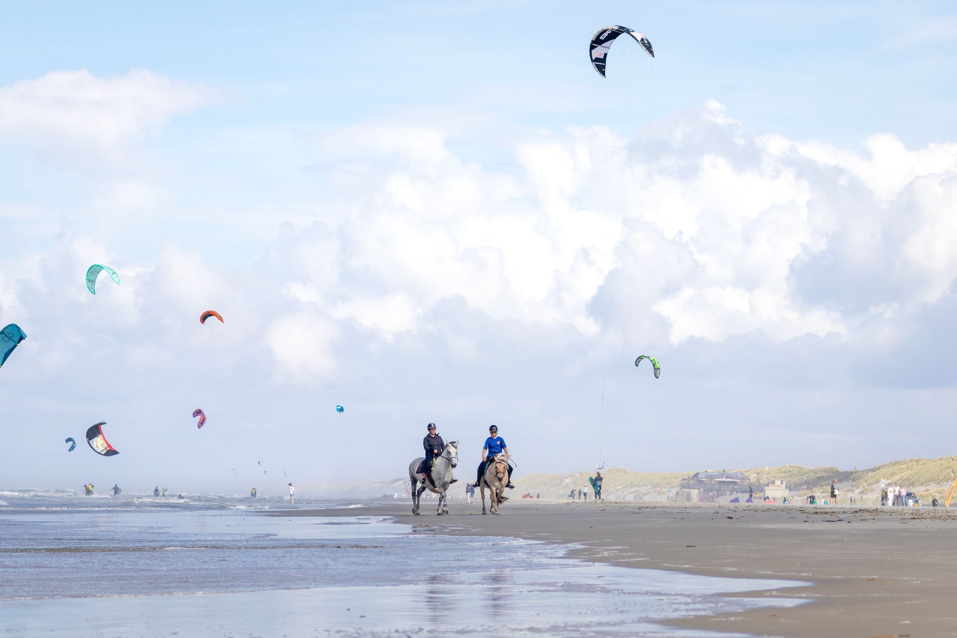 Twee mensen rijden te paard op een strand met kleurrijke vliegers in de lucht; het is een bewolkte dag.