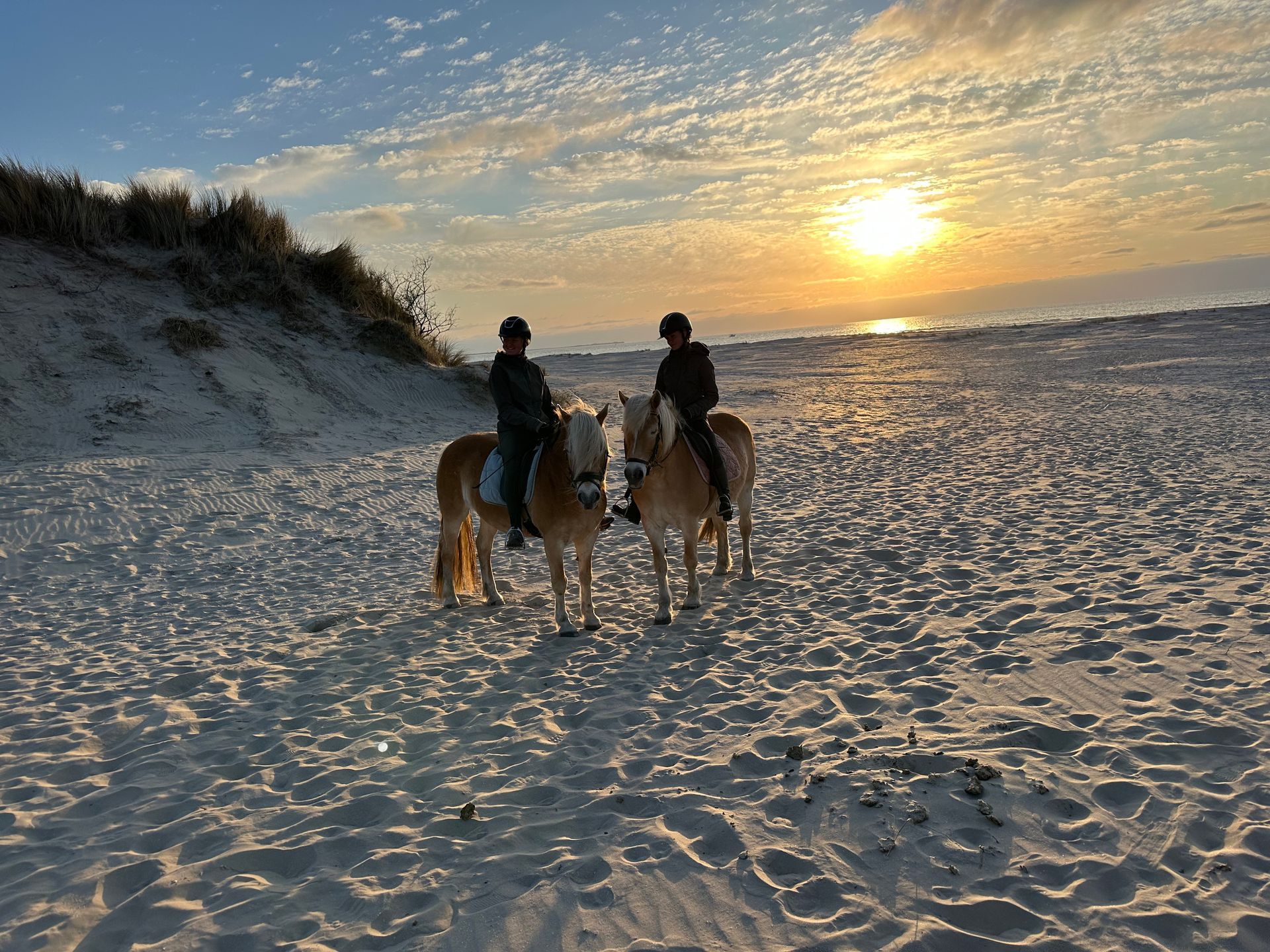 Two people are riding horses on the beach at sunset.