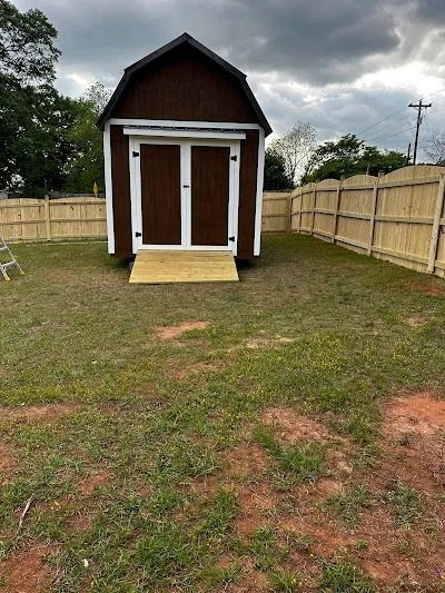 Brown shed with white trim, wooden ramp, and a surrounding wooden fence, in a grassy yard.