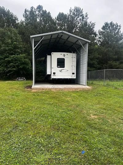 RV parked under a gray metal carport on a grassy area, with trees in the background.