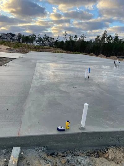 Newly poured concrete foundation with plumbing pipes, set against a cloudy sky.
