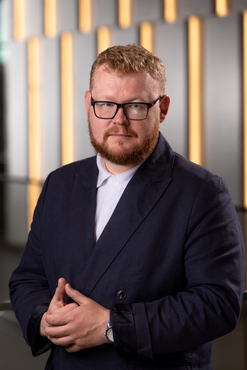 Portrait of a man in glasses and a navy suit standing with hands clasped against a softly lit background