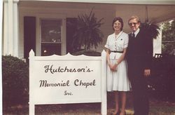 Man and woman standing beside a sign for Hutcheson's Memorial Chapel, a building with shutters and a porch, circa 1980s.