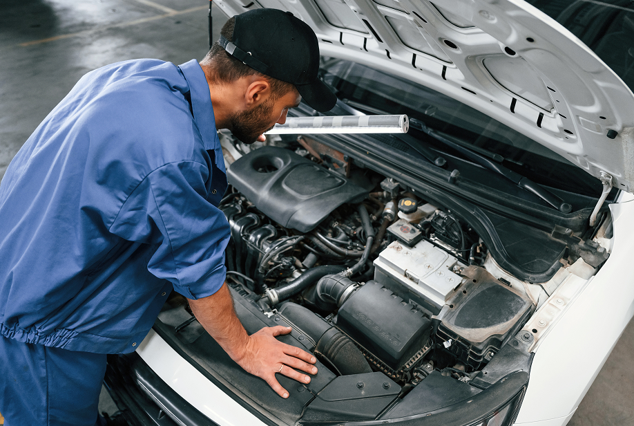 Mechanic in blue jumpsuit examines a car engine with a flashlight in a garage. | Lakeside Service Center