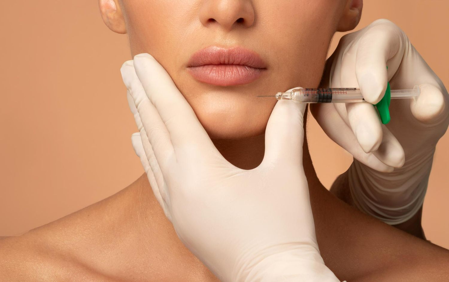 A gloved hand holds a person's chin while another hand prepares to administer a facial injection.
