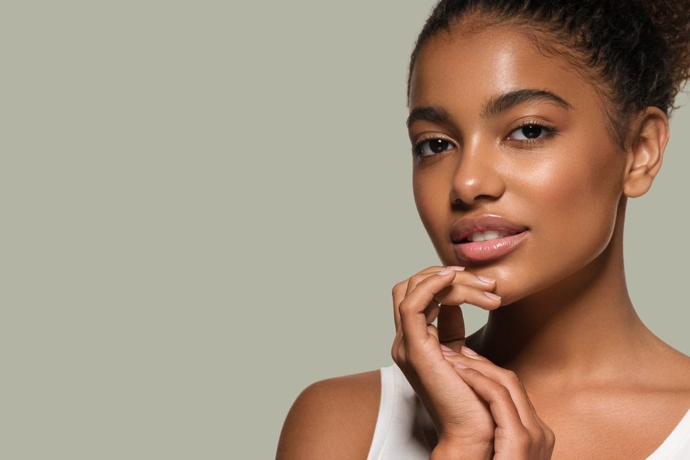 A person with glowing, healthy skin resting their chin on their hand against a soft, sage green background.