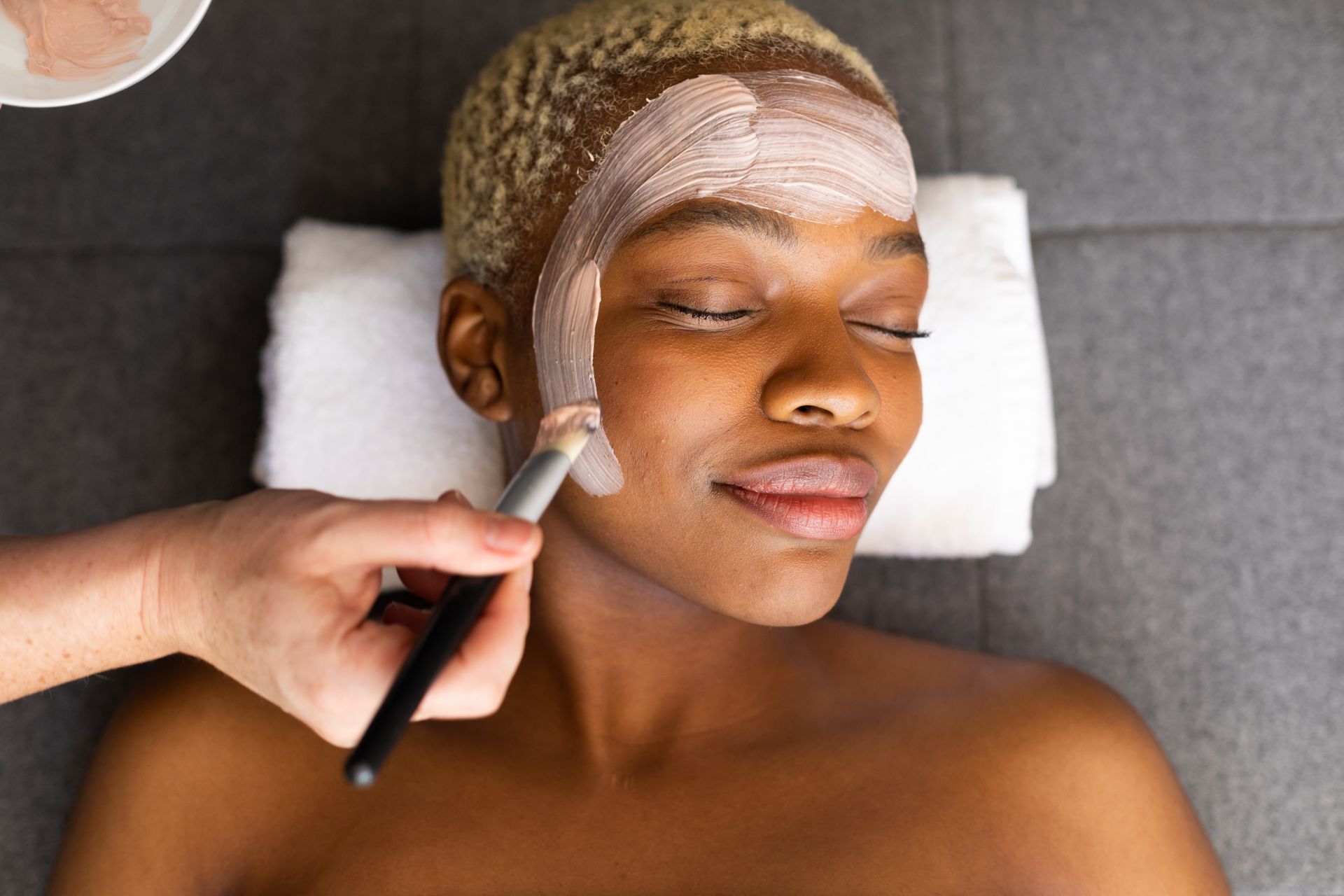 Person receiving a facial mask application with a brush while lying on a treatment bed