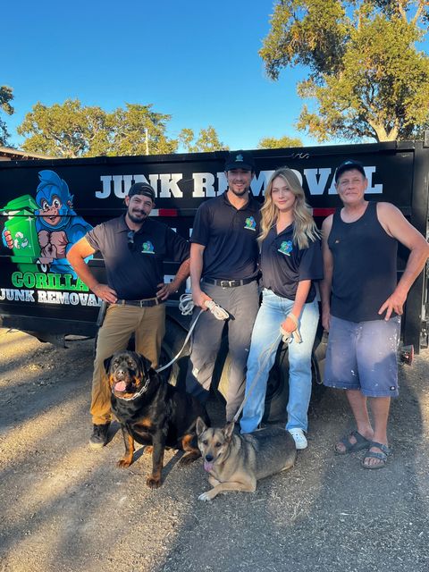 Four people and two dogs posing in front of a junk removal truck outdoors under a blue sky