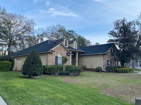Suburban brick house with a dark roof, front lawn, and trees under a partly cloudy sky