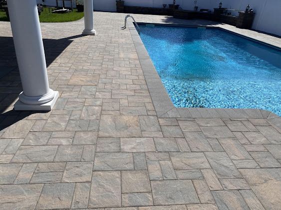 Stone pool deck beside a blue swimming pool with white columns and a grassy yard in the background