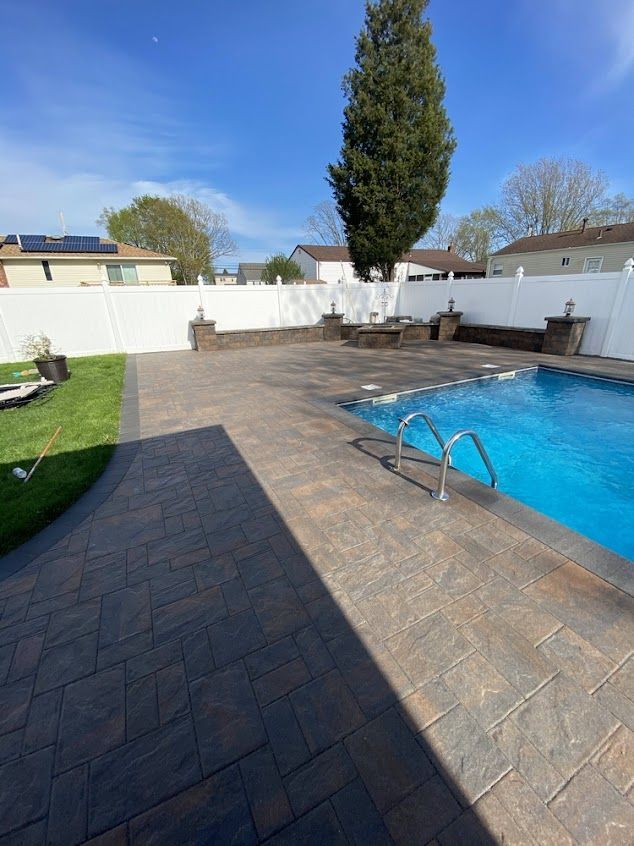 Backyard patio with swimming pool, paving stones, white fence, and tall tree under a blue sky