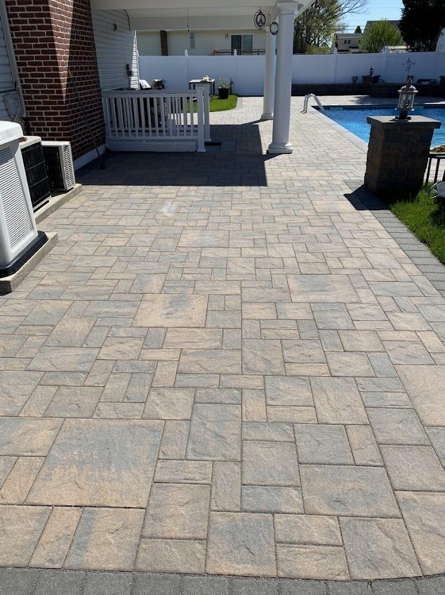 Stone paver patio leading to a covered porch, with white railing and a pool in the background.