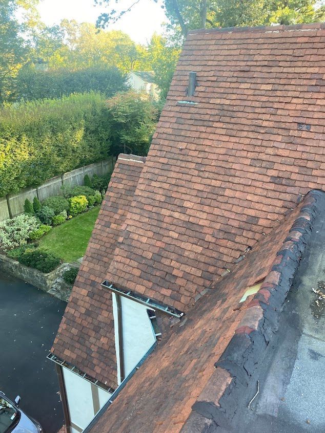 A steep red-tiled roof beside a leafy garden and driveway, viewed from above.