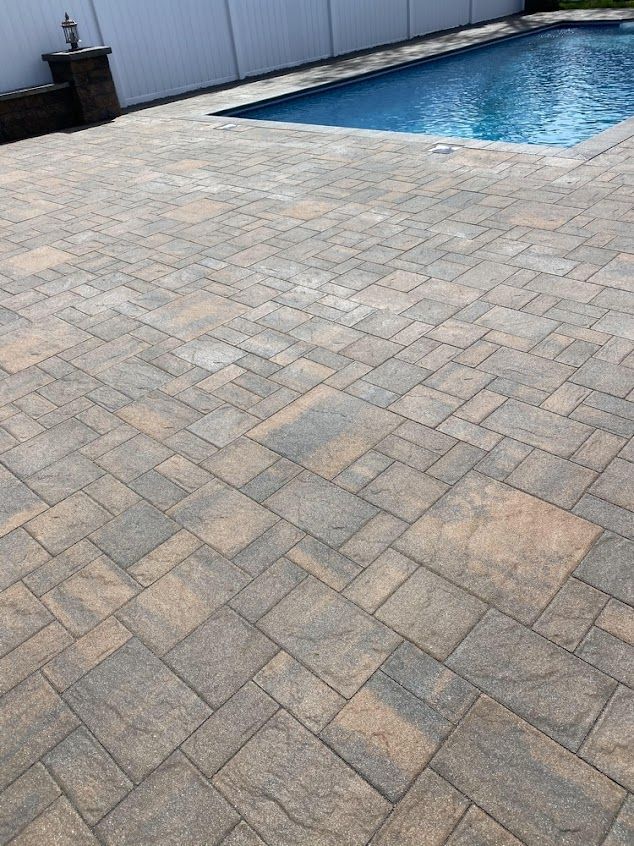 Paver patio beside a blue swimming pool and white fence, with a potted plant in the corner