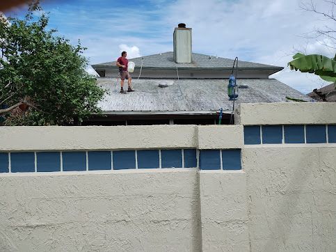 Workers on a rooftop repairing shingles beside a chimney, with a wall and blue sky in view